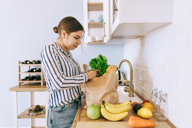 Joven mujer caucásica tomando verduras de una bolsa de papel en la cocina.