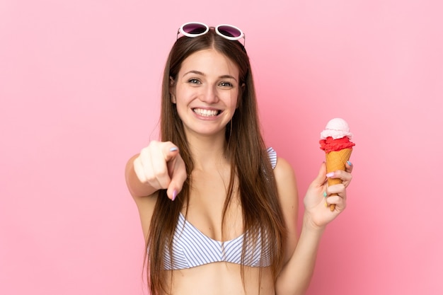 Joven mujer caucásica en traje de baño sosteniendo un helado de cucurucho aislado sobre fondo rosa le señala con el dedo con una expresión de confianza