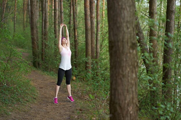 Joven mujer caucásica en traje de deporte estiramiento antes de correr en el bosque
