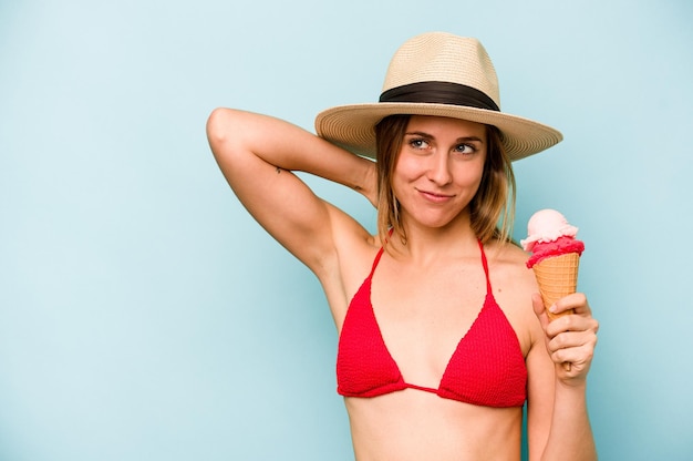 Joven mujer caucásica usando un bikini y sosteniendo un helado aislado en un fondo azul tocando la parte posterior de la cabeza pensando y tomando una decisión