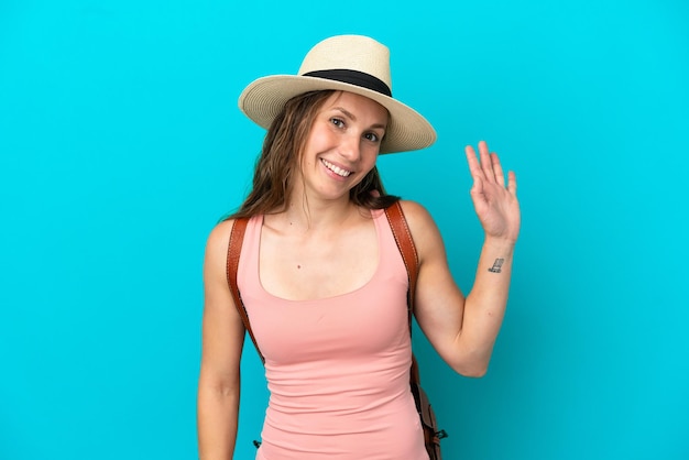 Joven mujer caucásica en vacaciones de verano aislado sobre fondo azul saludando con la mano con expresión feliz