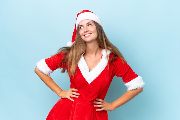 Joven mujer caucásica vestida como mamá noel aislada de fondo azul posando con los brazos en la cadera y sonriendo