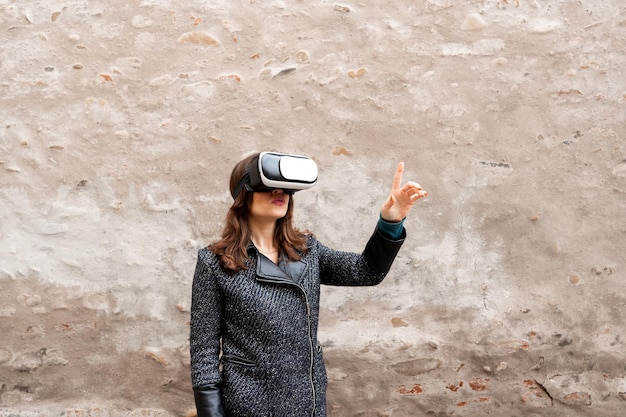 Joven mujer caucásica viendo por gafas de realidad virtual una pantalla digital y tocando con el dedo. Antiguo muro de fondo con espacio de copia.