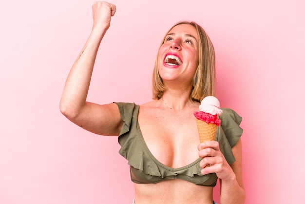 Joven mujer caucásica vistiendo un bikini y sosteniendo un helado aislado en un fondo rosa levantando el puño después de un concepto de ganador de la victoria