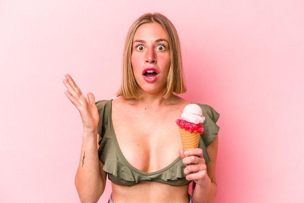 Joven mujer caucásica vistiendo un bikini y sosteniendo un helado aislado en un fondo rosado sorprendida y conmocionada