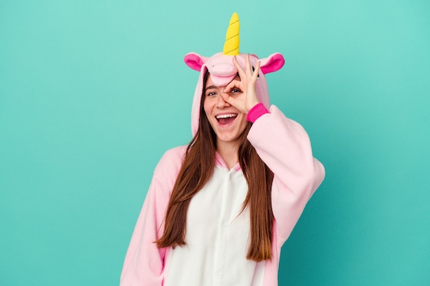 Joven mujer caucásica vistiendo un pijama de unicornio aislado sobre fondo azul emocionado manteniendo el gesto ok en el ojo.