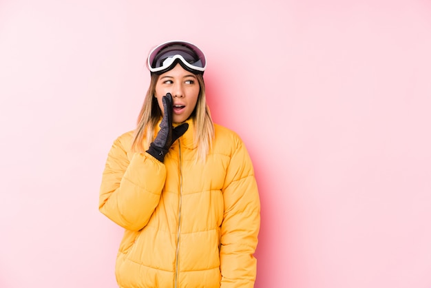 Joven mujer caucásica vistiendo ropa de esquí en una pared rosa está diciendo una noticia secreta de frenado en caliente y mirando a un lado
