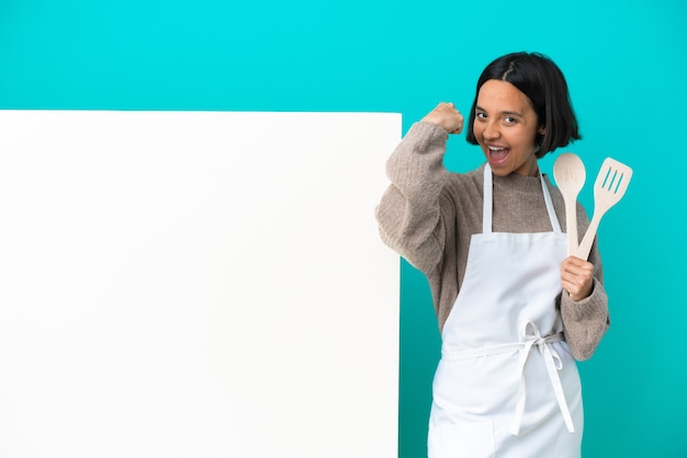 Joven mujer cocinera de raza mixta con un gran cartel aislado sobre fondo azul haciendo gesto fuerte
