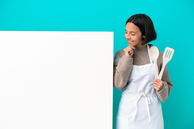 Joven mujer cocinera de raza mixta con un gran cartel aislado sobre fondo azul mirando hacia el lado y sonriendo