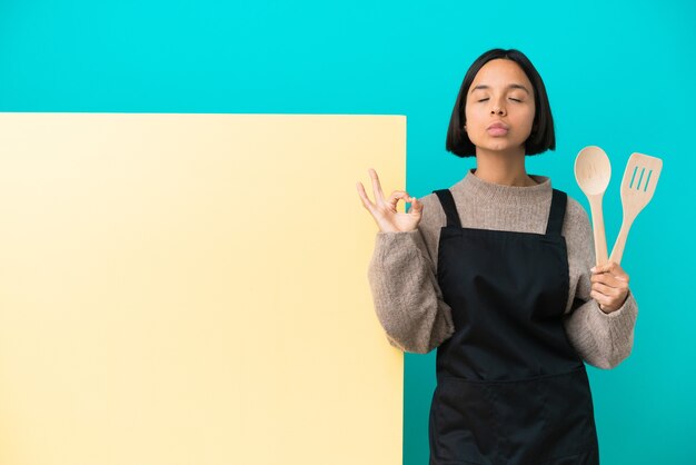 Joven mujer cocinera de raza mixta con un gran cartel aislado sobre fondo azul en pose zen