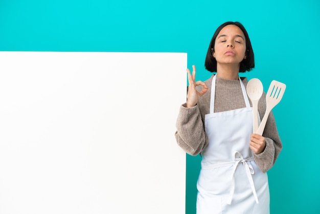 Joven mujer cocinera de raza mixta con un gran cartel aislado sobre fondo azul en pose zen