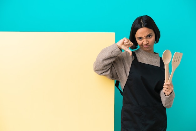 Joven mujer cocinera de raza mixta con un gran cartel aislado sobre fondo azul que muestra el pulgar hacia abajo con las dos manos