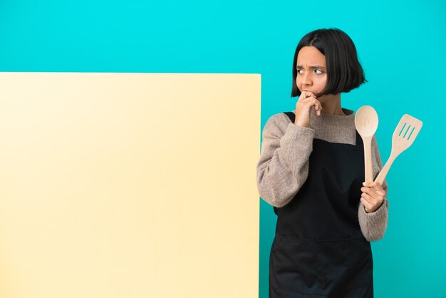 Joven mujer cocinera de raza mixta con un gran cartel aislado sobre fondo azul que tiene dudas y con expresión de la cara confusa