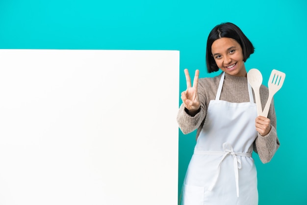 Joven mujer cocinera de raza mixta con un gran cartel aislado sobre fondo azul sonriendo y mostrando el signo de la victoria