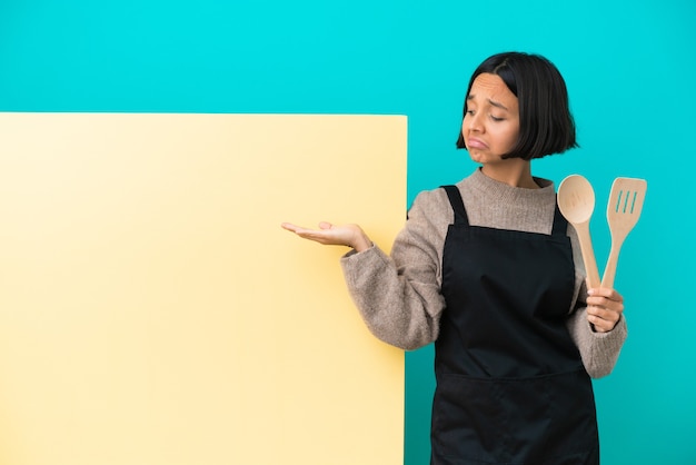 Joven mujer cocinera de raza mixta con un gran cartel aislado sobre fondo azul sosteniendo copyspace imaginario en la palma para insertar un anuncio