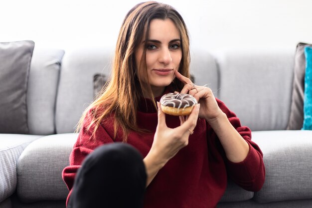 Joven mujer comiendo un donut sentado en la alfombra cerca de un sofá