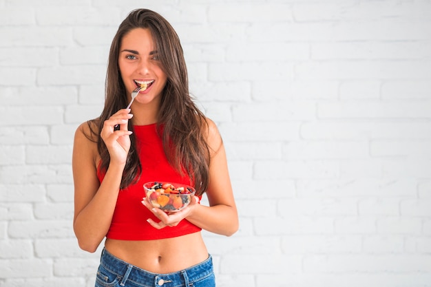 Joven mujer comiendo ensalada de frutas con tenedor de pie contra la pared blanca