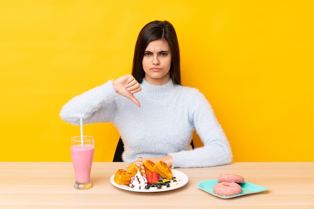 Joven mujer comiendo gofres y batidos en una mesa sobre la pared amarilla mostrando el pulgar hacia abajo signo