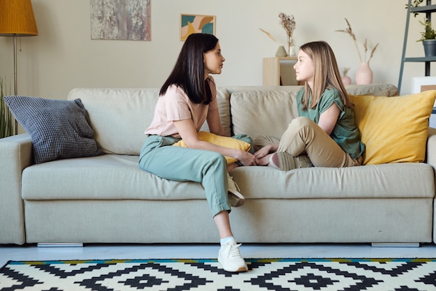 Joven mujer contemporánea y su hija en ropa casual sentados en el sofá uno frente al otro mientras discuten los planes para el día