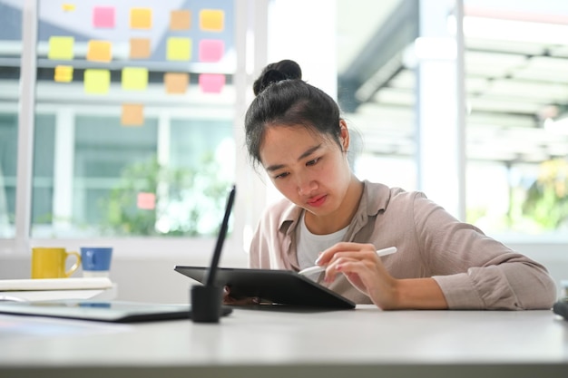 Joven mujer creativa concentrada trabajando en tableta digital mientras está sentada en la oficina