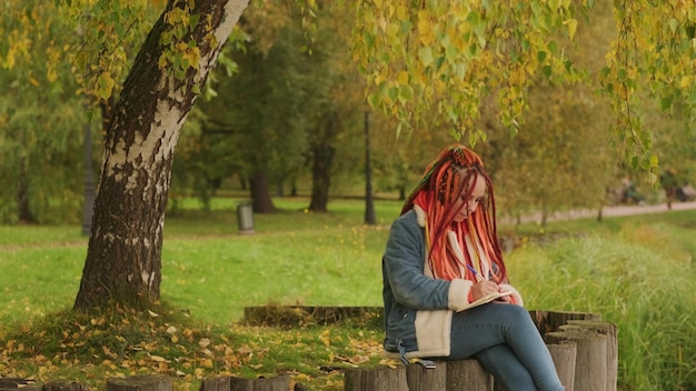 Joven mujer creativa con rastas dibujando escritura en un cuaderno sentado en tocones cerca del lago en un parque