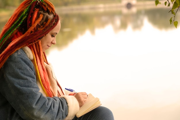 Joven mujer creativa con rastas dibujando escritura en un cuaderno sentado en tocones cerca del lago en un parque