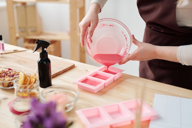 Joven mujer creativa vertiendo masa de jabón líquido rosa del tazón en moldes de silicona mientras está de pie junto al lugar de trabajo