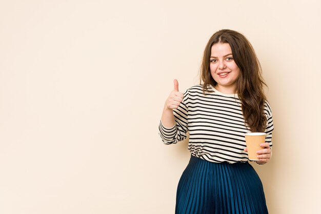 Joven mujer con curvas sosteniendo un café sonriendo y levantando el pulgar