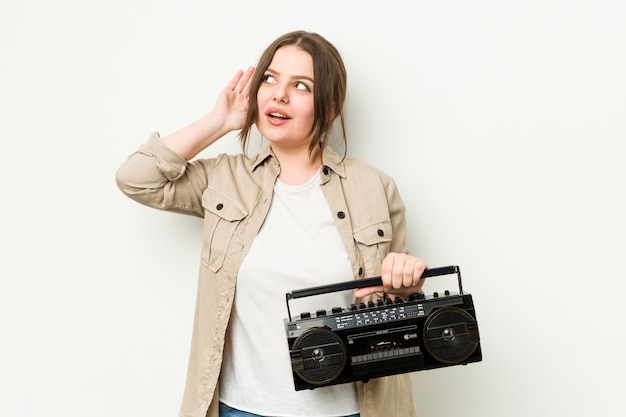 Joven mujer con curvas sosteniendo una radio retro tratando de escuchar un chisme.