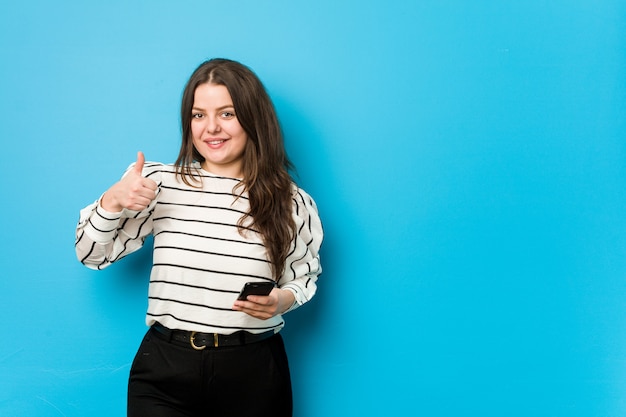 Joven mujer con curvas sosteniendo un teléfono sonriendo y levantando el pulgar