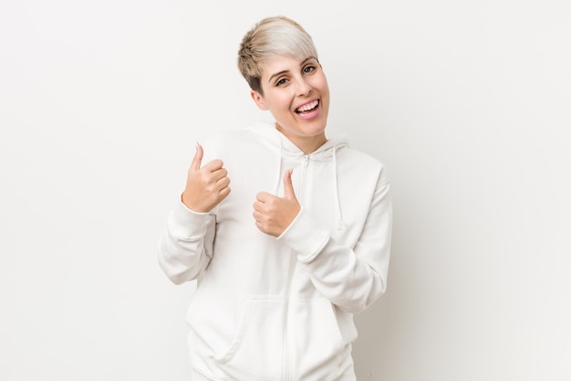 Joven mujer con curvas con una sudadera con capucha blanca levantando ambos pulgares, sonriente y confiado.