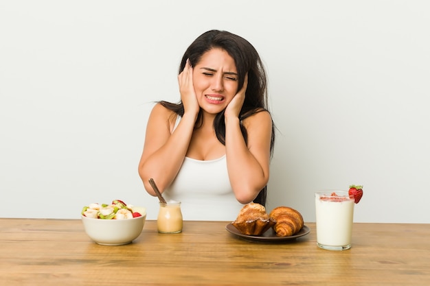 Joven mujer con curvas tomando un desayuno cubriendo las orejas con las manos.