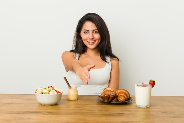 Joven mujer con curvas tomando un desayuno estirando la mano a la cámara en gesto de saludo