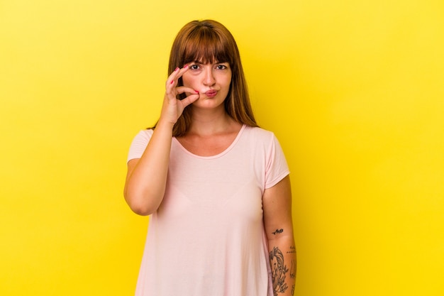 Joven mujer curvilínea caucásica aislada sobre fondo amarillo con los dedos en los labios guardando un secreto.