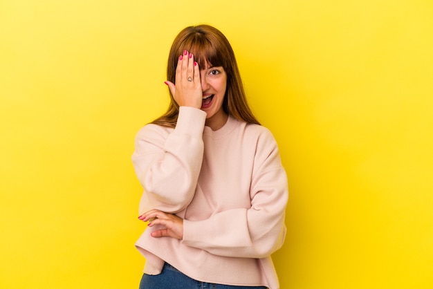 Joven mujer curvilínea caucásica aislada sobre fondo amarillo divirtiéndose cubriendo la mitad de la cara con la palma.