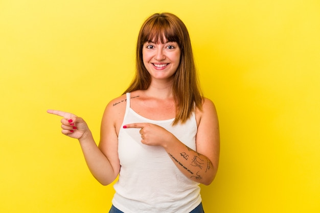 Joven mujer curvilínea caucásica aislada sobre fondo amarillo emocionada apuntando con los dedos índices.