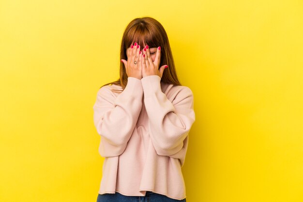 Joven mujer curvilínea caucásica aislada sobre fondo amarillo parpadea a la cámara a través de los dedos, avergonzado cubriendo la cara.