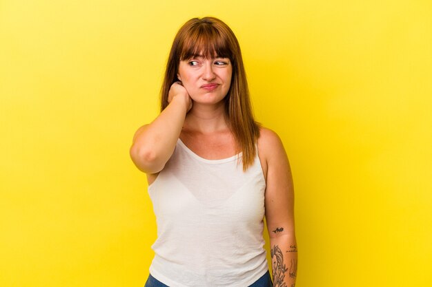 Joven mujer curvilínea caucásica aislada sobre fondo amarillo tocando la parte posterior de la cabeza, pensando y haciendo una elección.