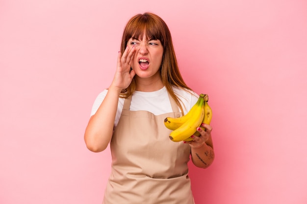 Joven mujer curvilínea caucásica cocinando en casa sosteniendo plátanos aislados sobre fondo rosa gritando y sosteniendo la palma cerca de la boca abierta.