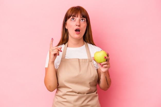 Joven mujer curvilínea caucásica cocinar en casa sosteniendo una manzana aislada sobre fondo rosa apuntando al revés con la boca abierta.