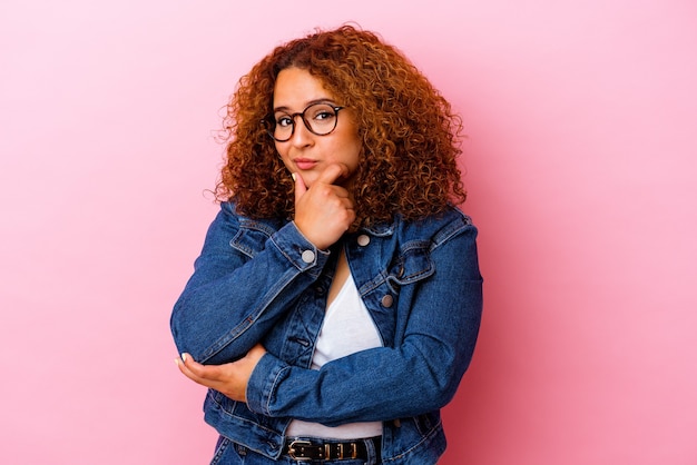 Joven mujer curvilínea latina aislada en pared rosa sospechosa, incierta, examinándote.