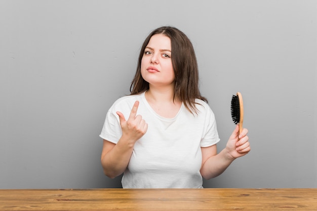 Joven mujer curvilínea de talla grande sosteniendo un cepillo para el cabello apuntando con el dedo como si invitara a acercarse.