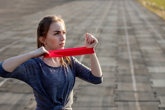 Joven mujer delgada en ropa deportiva haciendo sentadillas ejercicio con banda elástica en una pista del estadio recubierto de negro