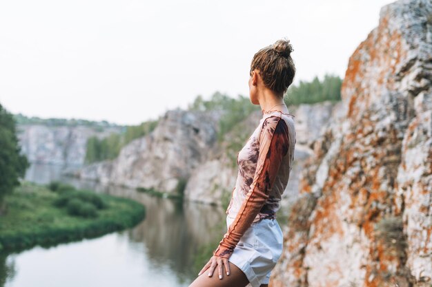 Joven mujer delgada con ropa informal mira la hermosa vista de las montañas y los tranquilos viajes locales por el río