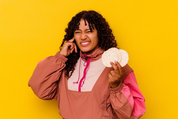 Joven mujer de deporte de raza mixta sosteniendo un pastel de arroz aislado en amarillo que cubre las orejas con las manos.