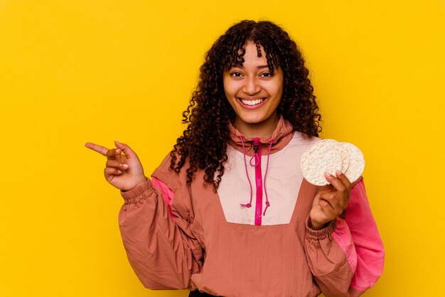 Joven mujer de deporte de raza mixta sosteniendo un pastel de arroz aislado sobre fondo amarillo sonriendo y apuntando a un lado, mostrando algo en el espacio en blanco.