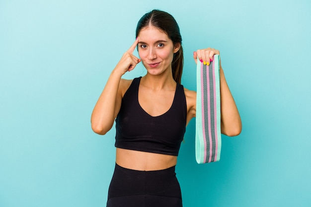 Foto joven mujer deportiva caucásica sosteniendo bandas elásticas aisladas sobre fondo azul señalando la sien con el dedo, pensando, centrado en una tarea.