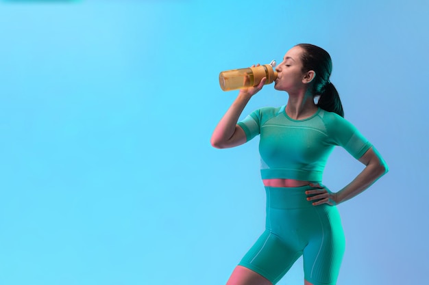 Joven mujer deportiva entrenando y bebiendo agua, aislado sobre fondo de estudio degradado en luz de neón