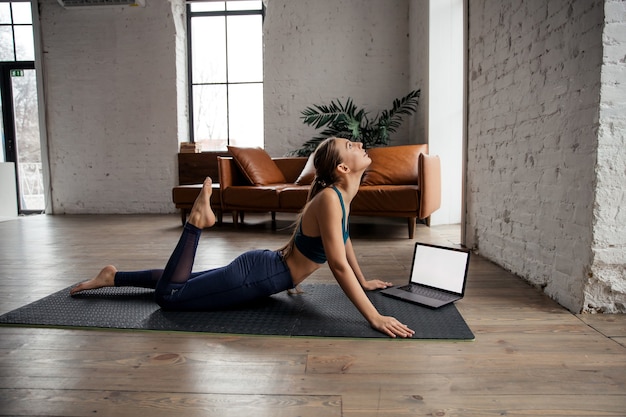 Joven mujer deportiva practicando yoga y estirando el cuerpo en casa usando la computadora portátil para clases en línea o tutoriales virtuales. Foto de alta calidad