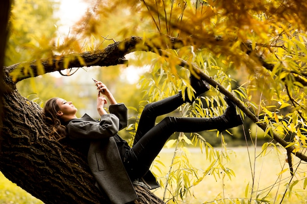 Joven mujer descansando en el árbol con teléfono móvil